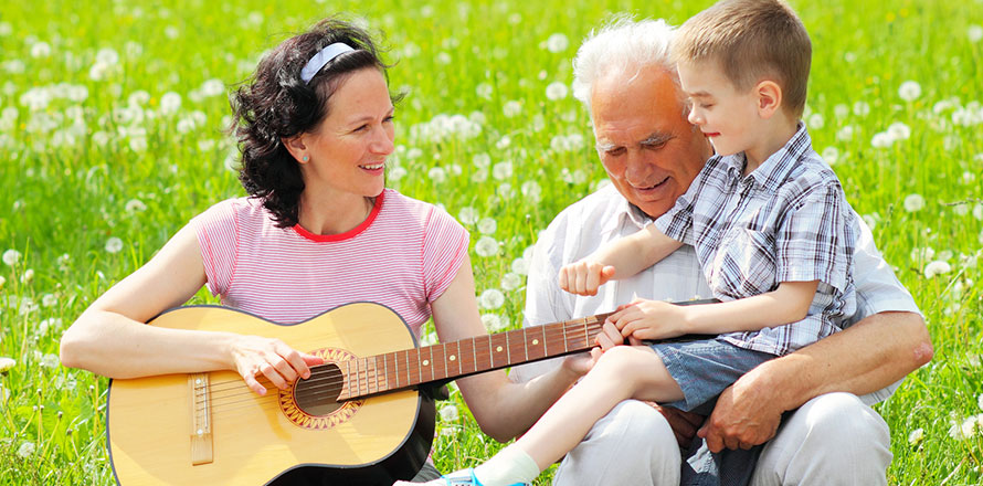 Mutter mit Gitarre auf einer Wiese, rechts daneben Großvater mit kleinem Jungen auf dem Schoß